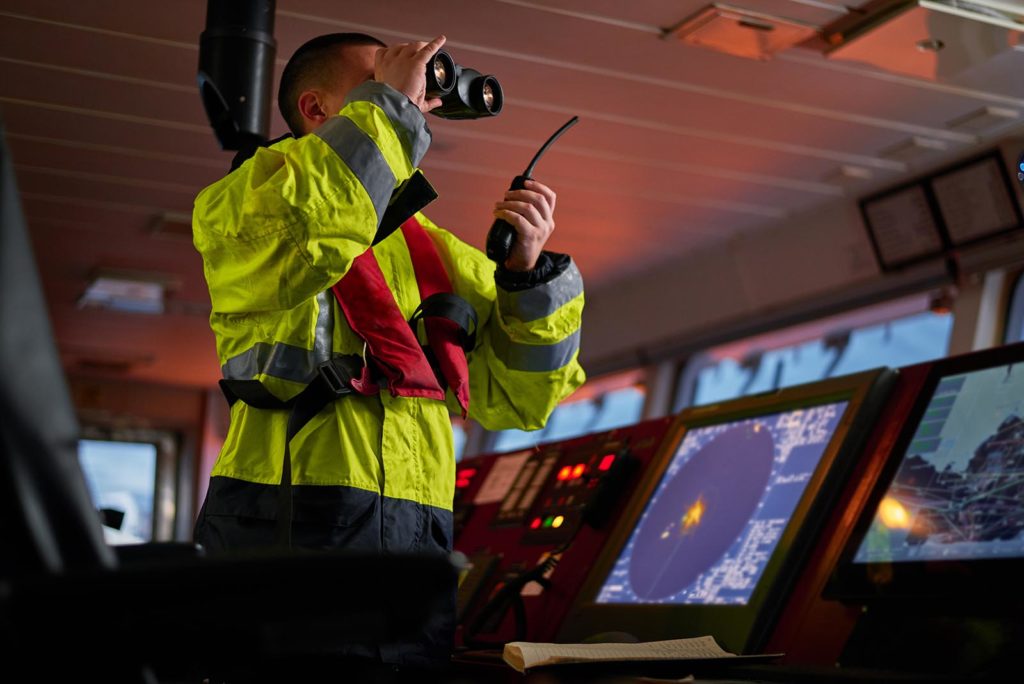 Navigator of ship crew performing daily duties with VHF radio, binoculars, logbook, standing nearby to ECDIS and radar station on board of modern ship